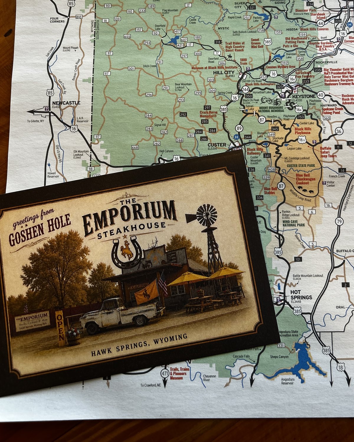 A Greetings from Goshen Hole postcard of The Emporium Steakhouse, lying on a Black Hills and Badlands road map